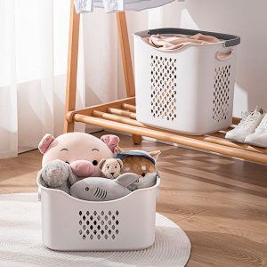 Giỏ nhựa đựng đồ chơi và thú bông đặt trên sàn cạnh kệ gỗ.