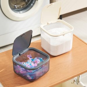 Hai hộp đựng nhựa có nắp, một đựng viên giặt, một đựng bột giặt, đặt trên bàn cạnh máy giặt.