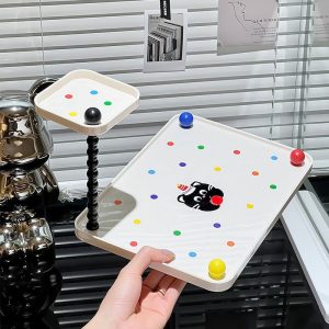 Kệ đựng đồ hai tầng với họa tiết mèo đen và chấm màu, có bi chặn ở các góc.