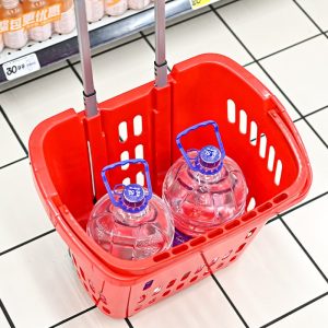 Giỏ kéo màu đỏ với tay kéo và hai bình nước lớn bên trong, đặt trên sàn siêu thị.
