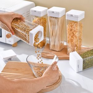 Hộp nhựa PP vuông kín khí đựng thực phẩm khô trong suốt tiện lợi Đổ hạt từ hộp vào bát, nhiều hộp đựng thực phẩm trên bàn bếp.