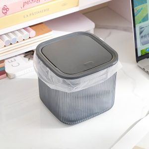 Hộp đựng nhựa màu xám khói, có lót túi rác nhỏ, đặt trên bàn làm việc.