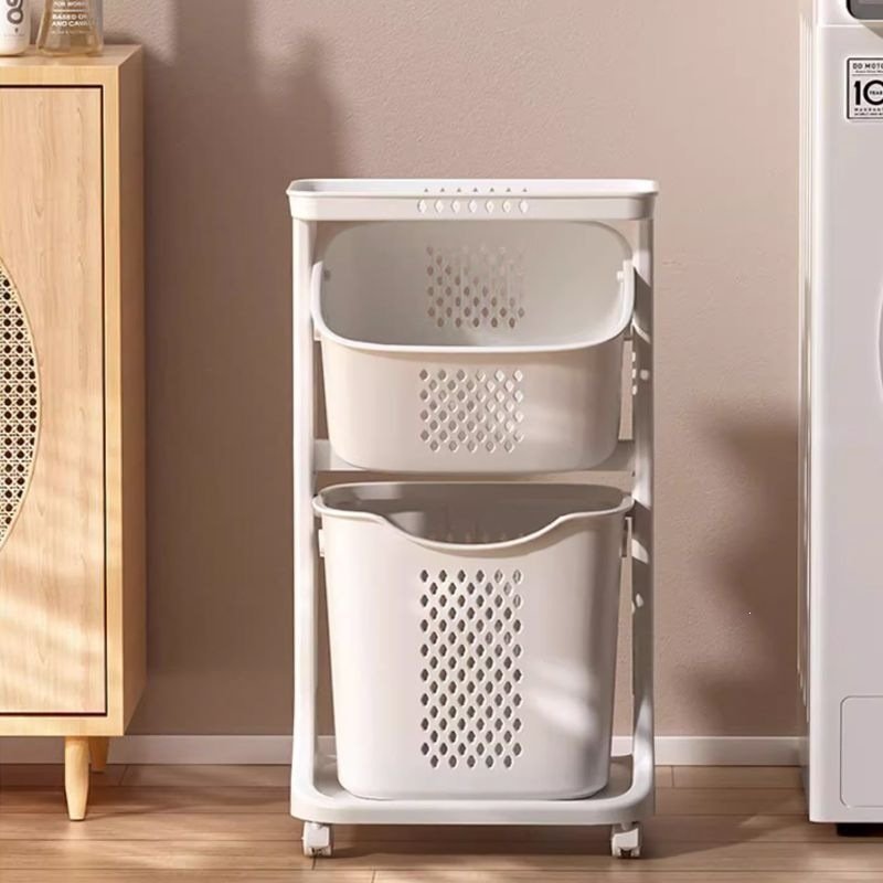 Giá để đồ nhựa PP 3 tầng có bánh xe đựng quần áo bẩn đa năng Một giỏ đựng quần áo 2 tầng màu trắng, đặt cạnh tủ gỗ và máy giặt.