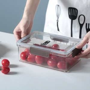 Một người đang cầm hộp nhựa trong suốt đựng cà chua bi trên bàn.