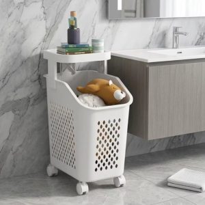 Giỏ đựng quần áo trắng đặt cạnh bồn rửa mặt, phía trên để sách và đồ dùng nhỏ